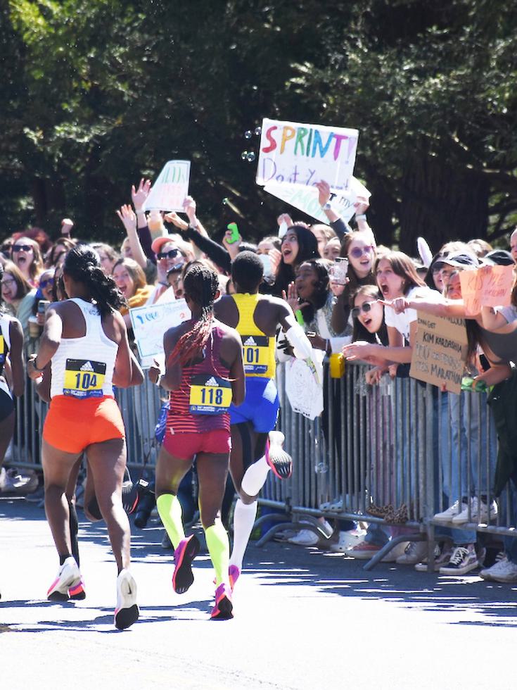 Students standing behind a barricade, holding signs and cheering marathon runners in the foreground