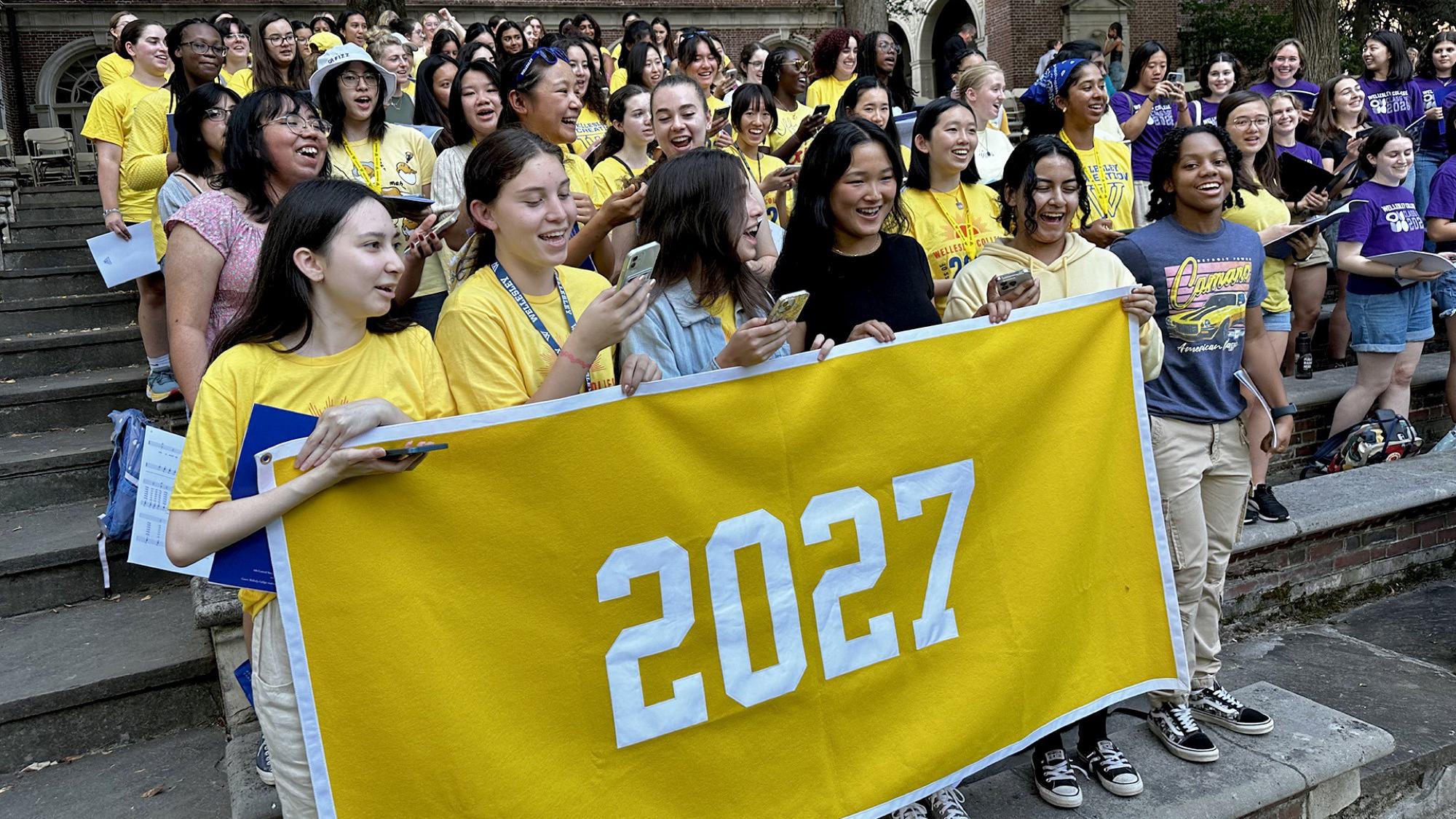 Members of the class of 2027 on steps, clad in yellow, holding a yellow class year banner, singing.