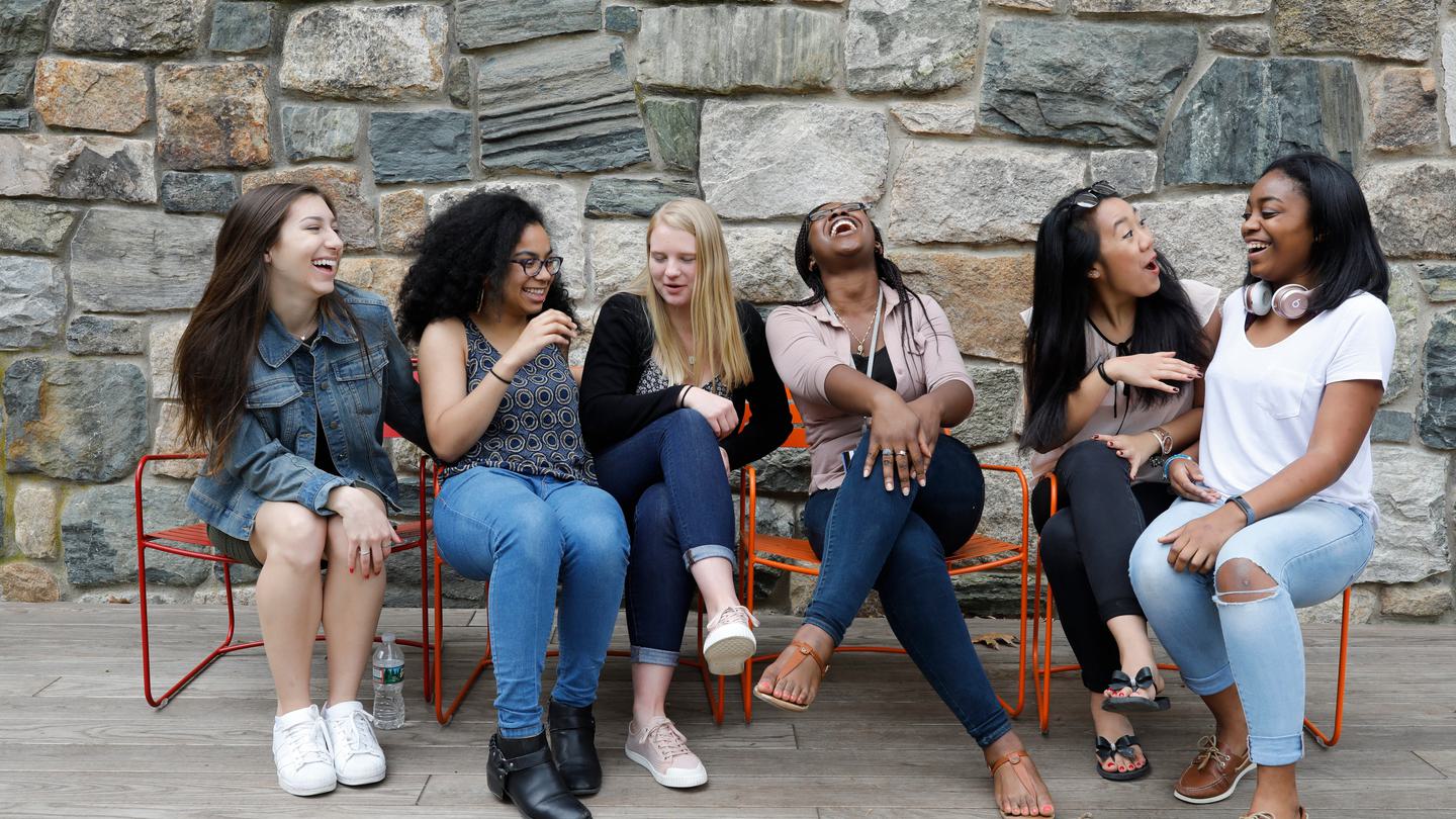 Six friends sit on a bench laughing in front of a stone building.