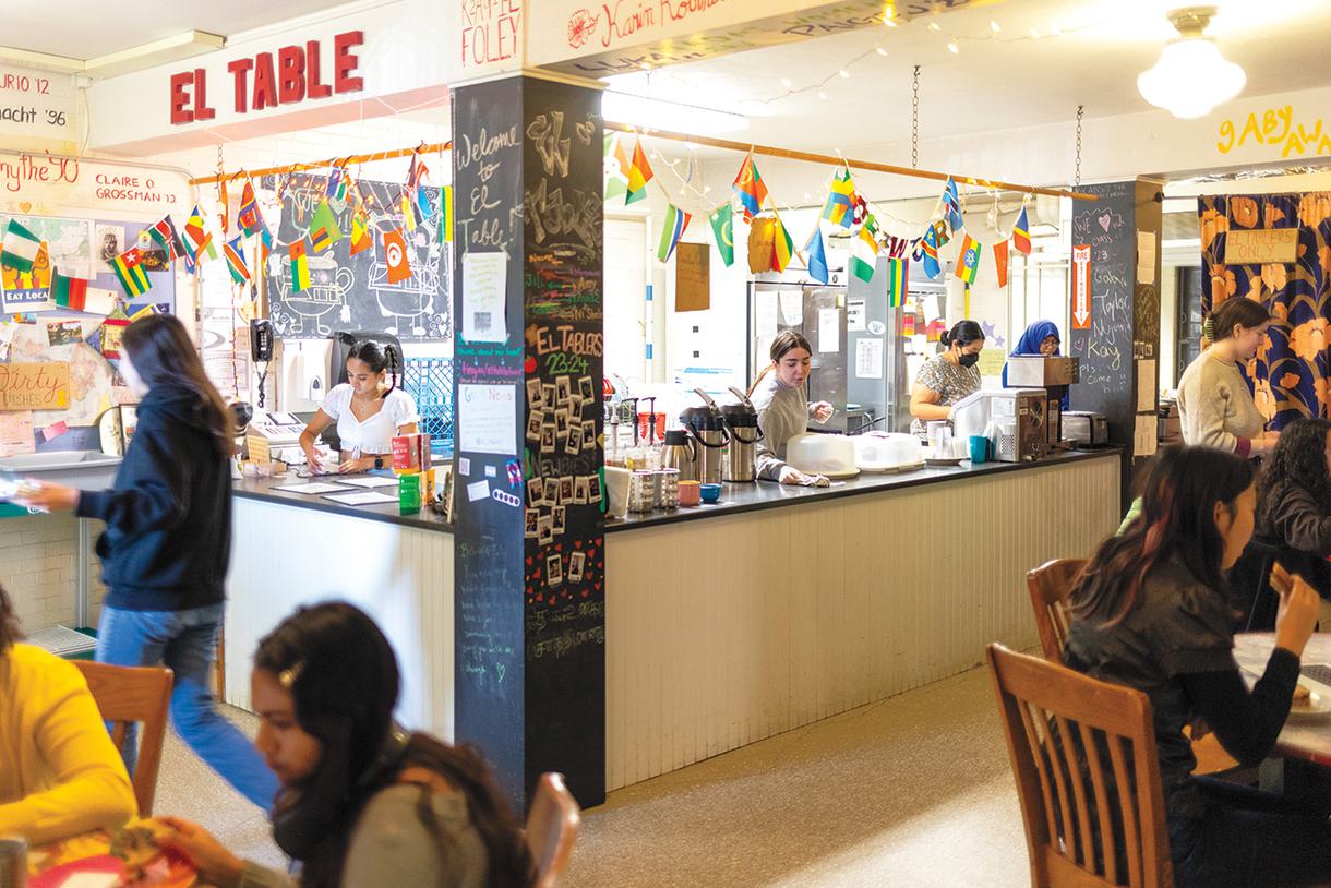 Four students work behind the counter at El Table while others munch on sandwiches at tables.
