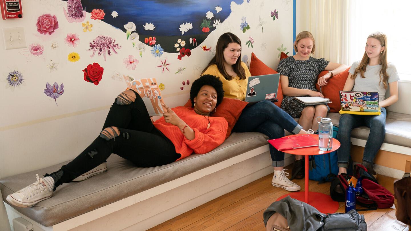 Four students hang out in a room with a colorful floral mural on the wall.