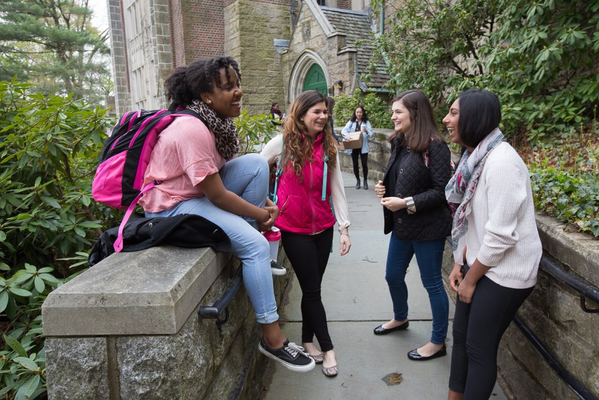 Students stand on a ramp, chatting. One sits on the wall.