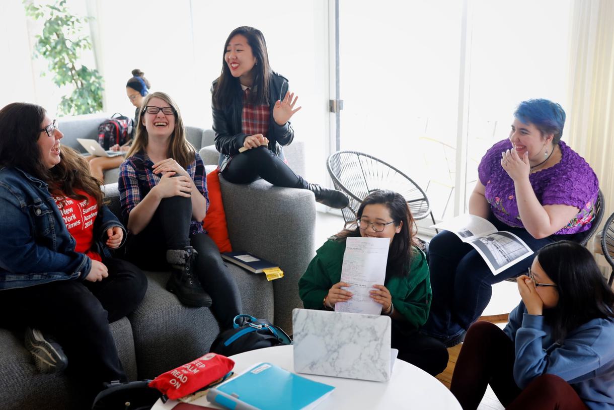 A group of students sitting on a sofa and chairs laugh while gathered around a table.