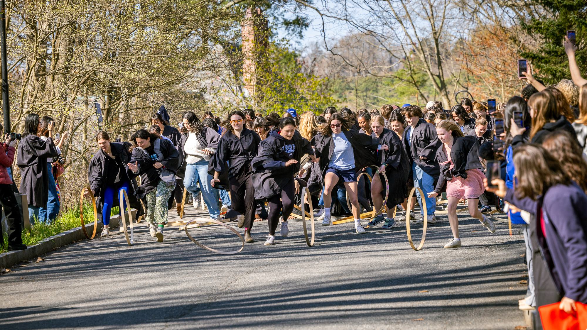 Seniors wearing graduation robes rolling wooden hoops down Tupelo Lane