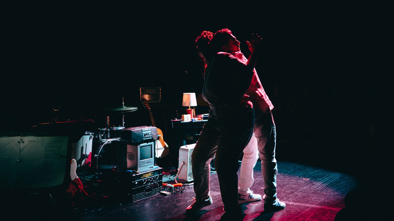 A woman and man dance back to back on a stage with their arms entwined. The are engulfed in shadows.