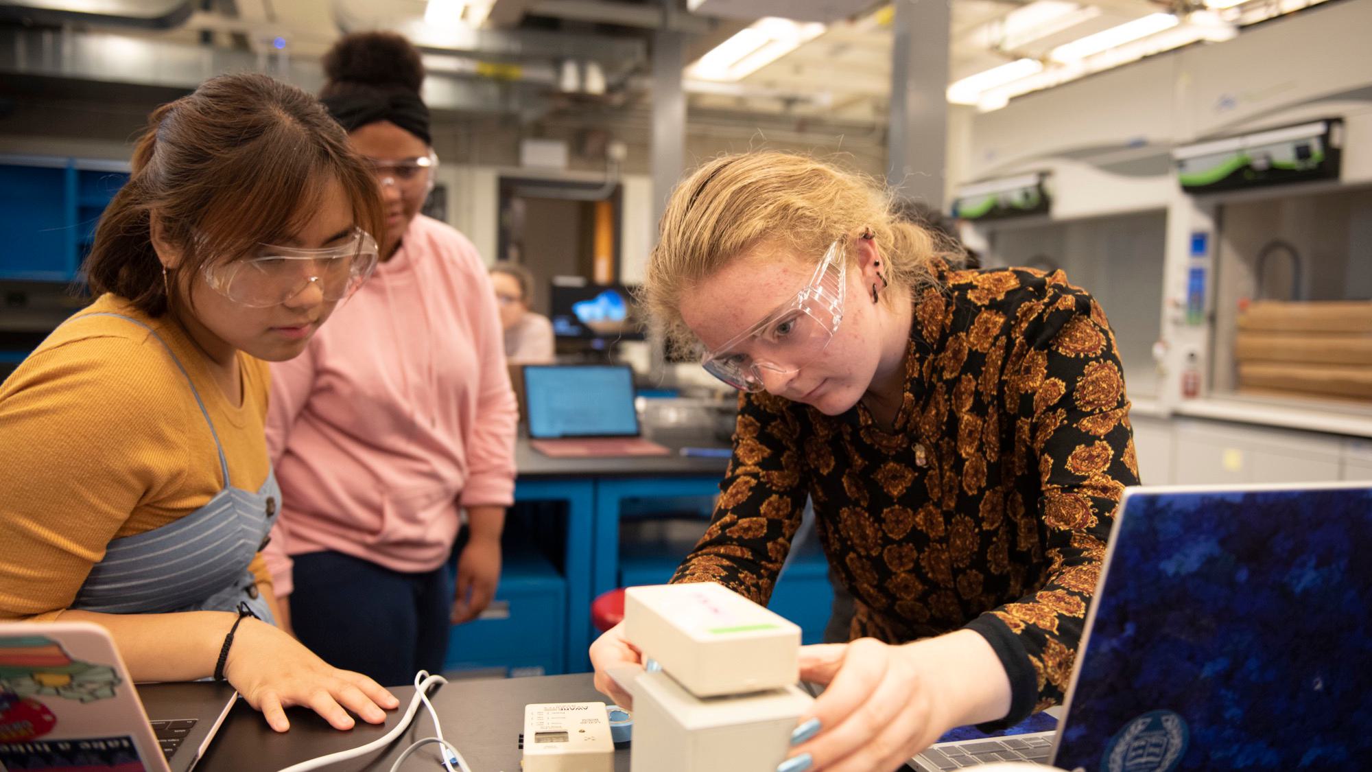 Students wearing safety goggles in a laboratory classroom use a piece of lab technology together.