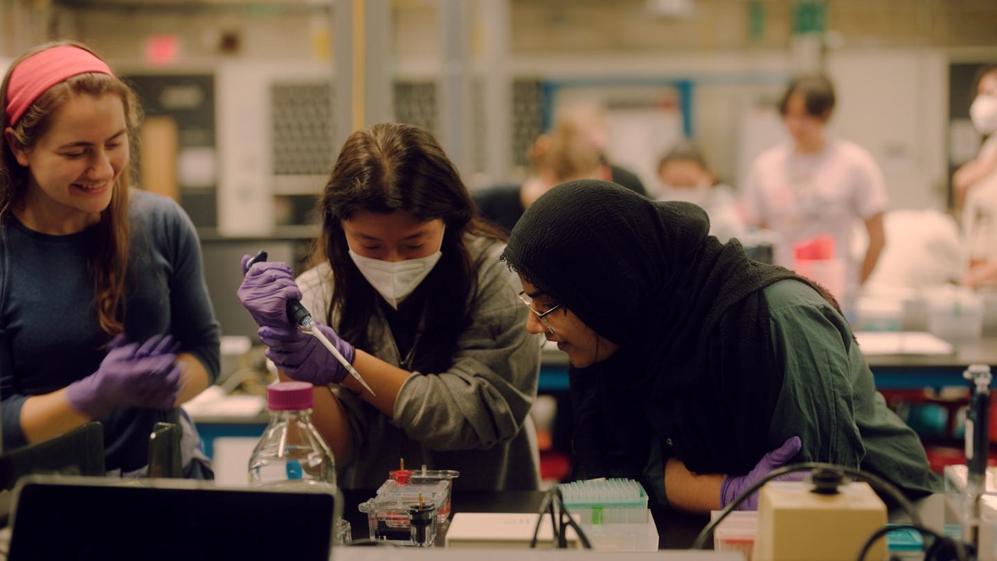 Two students observe a third pipetting into a gel in a science lab.