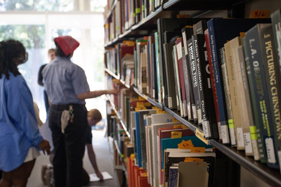 A closeup shot of a library bookshelf. Two students are browsing the shelf in the background.