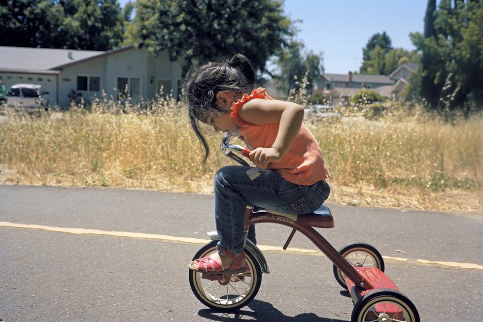 A child riding a tricycle on a street looks down at her feet on the pedals.