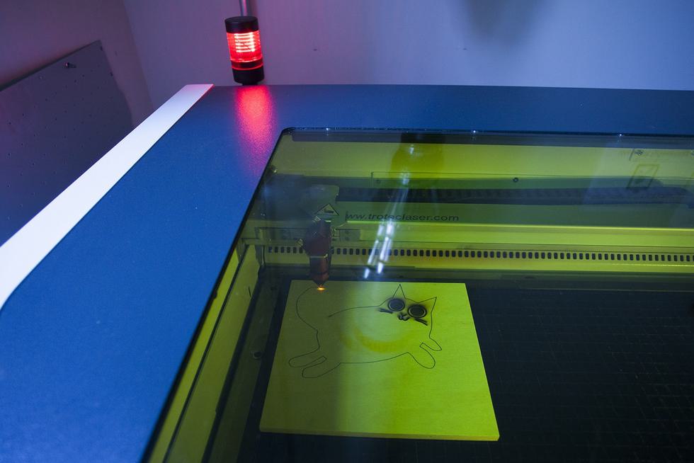 A laser cutter imprints the Art Department logo, a black cat, into a piece of wood.