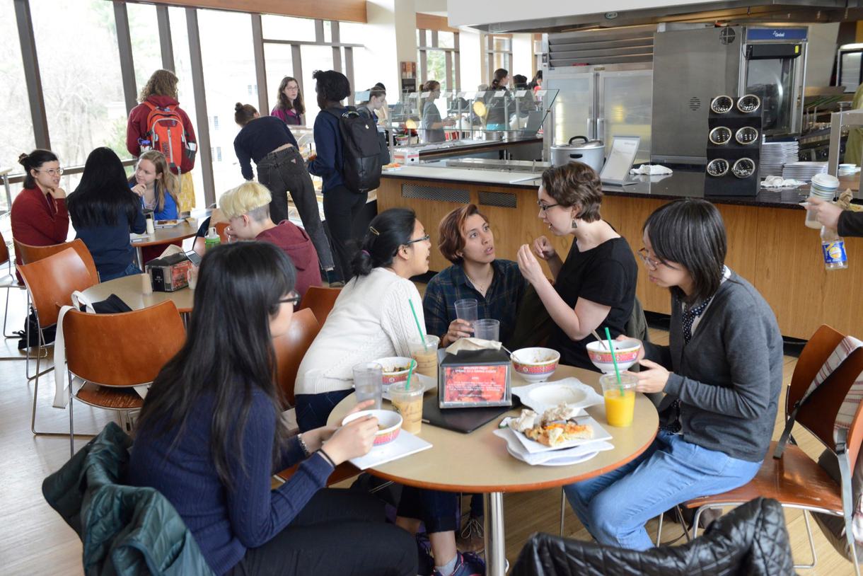 Groups of students eat around small round tables.