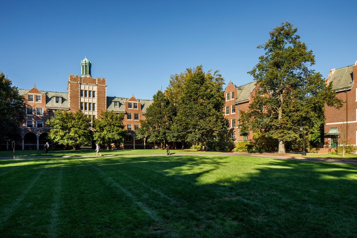 Students cross the grassy quad in front of a brick building with open archways.