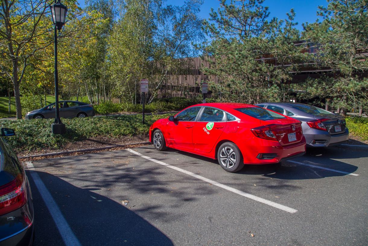 Red Civic with the Zipcar logo on it parked in a small outdoor parking lot.