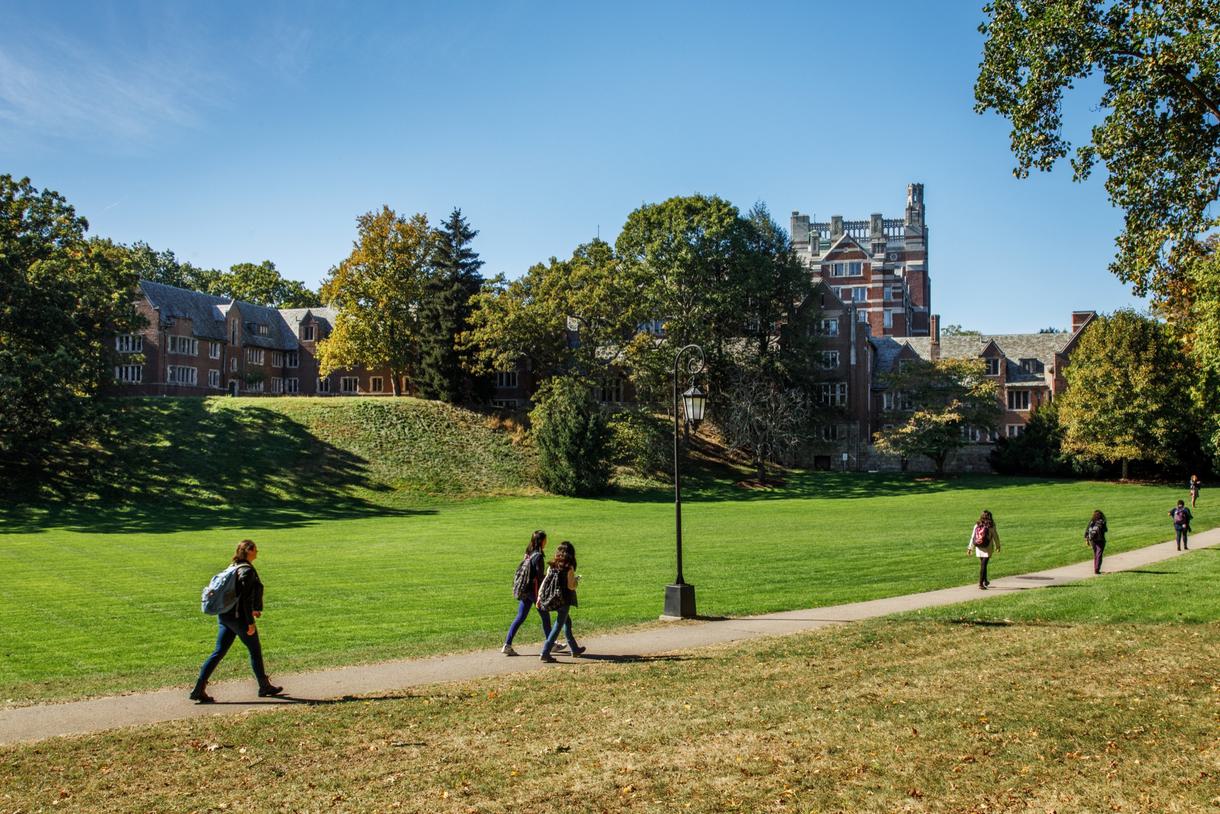 Brick buildings in the background against a blue sky. Students walking to class across a grassy lawn in the foreground.