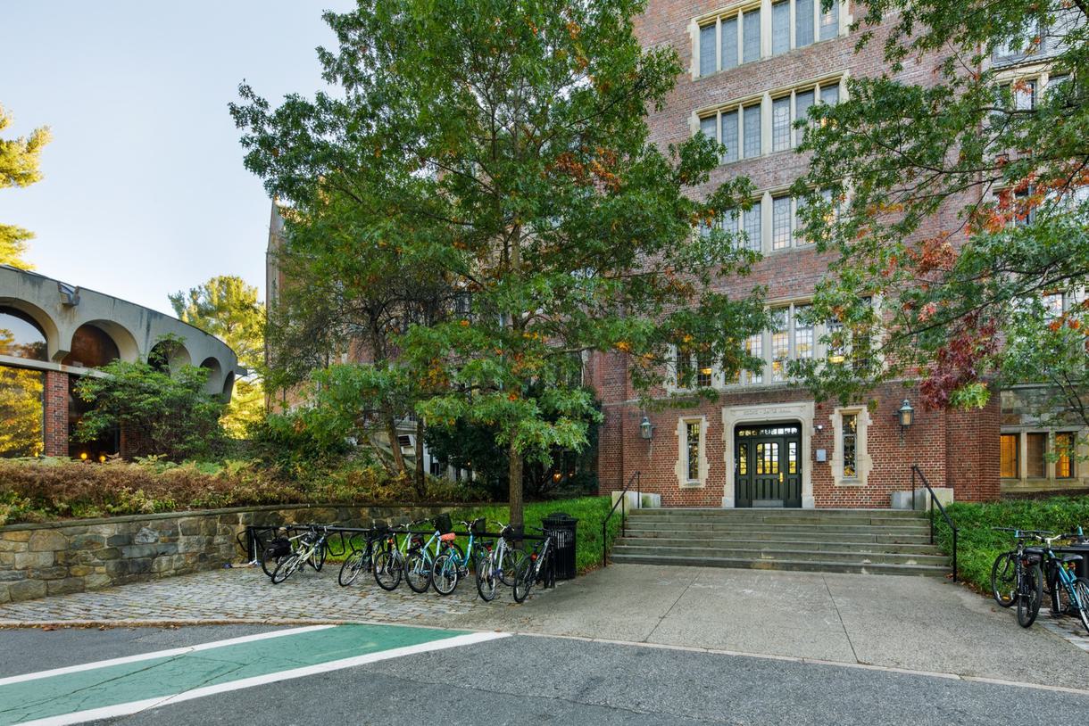 A brick building with stone steps is covered by trees. There are parked bicycles in the foreground.