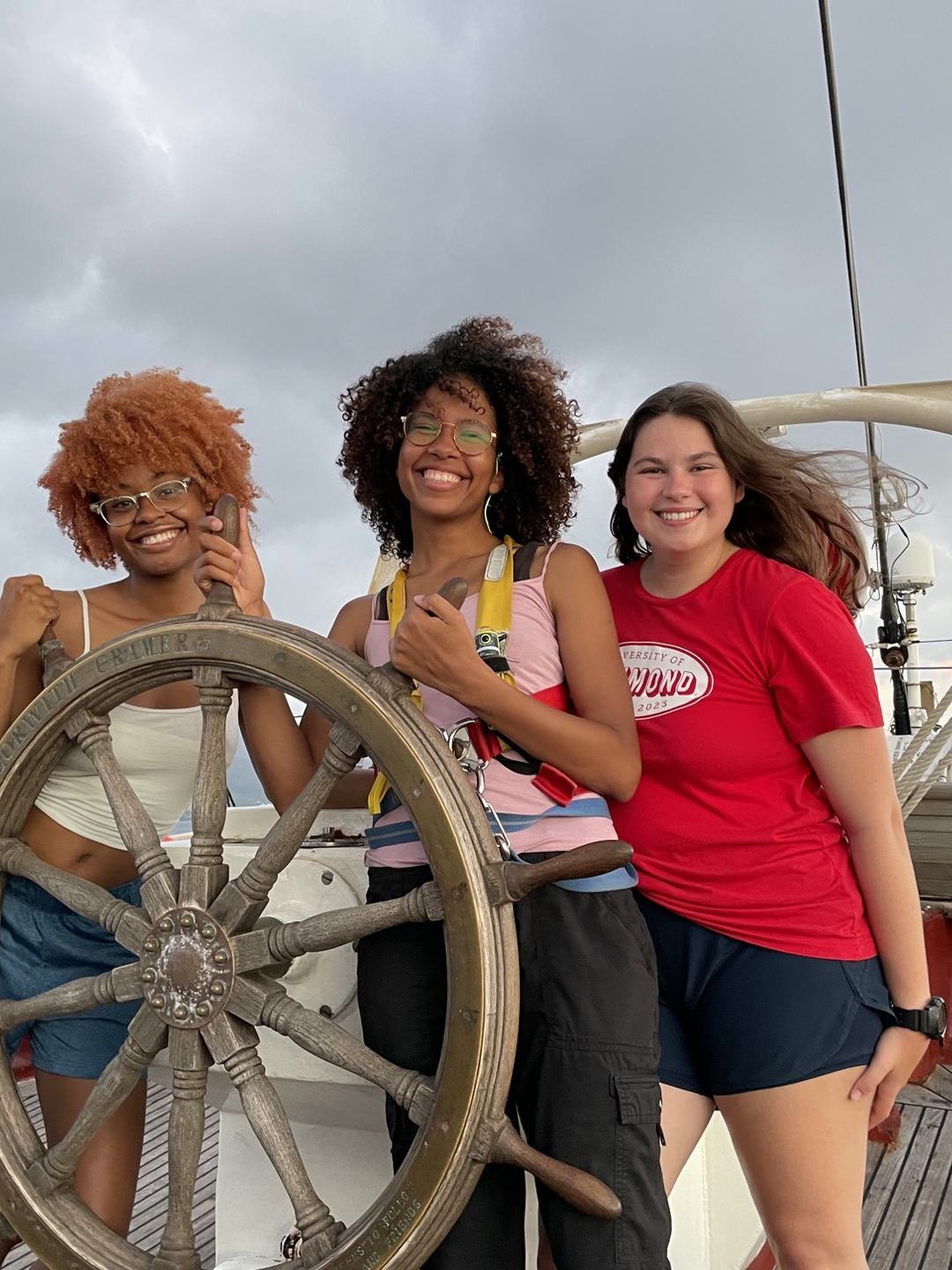 Three students pose, smiling at the helm of a ship.