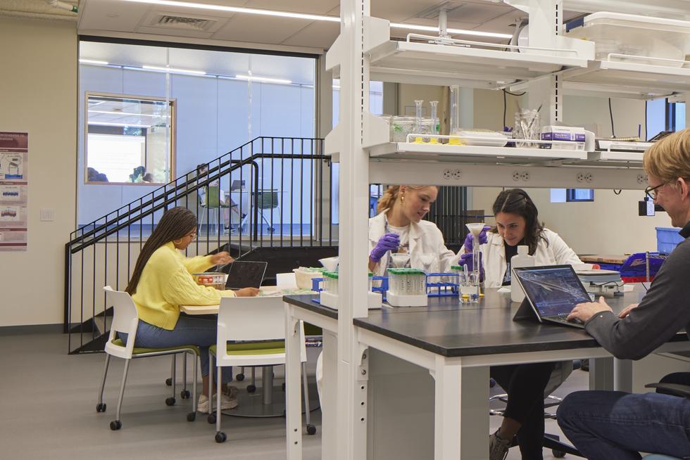 Students and a professor sit at lab tables. The professor looks at their laptop, the students examine a beaker. Another student looks at their laptop.