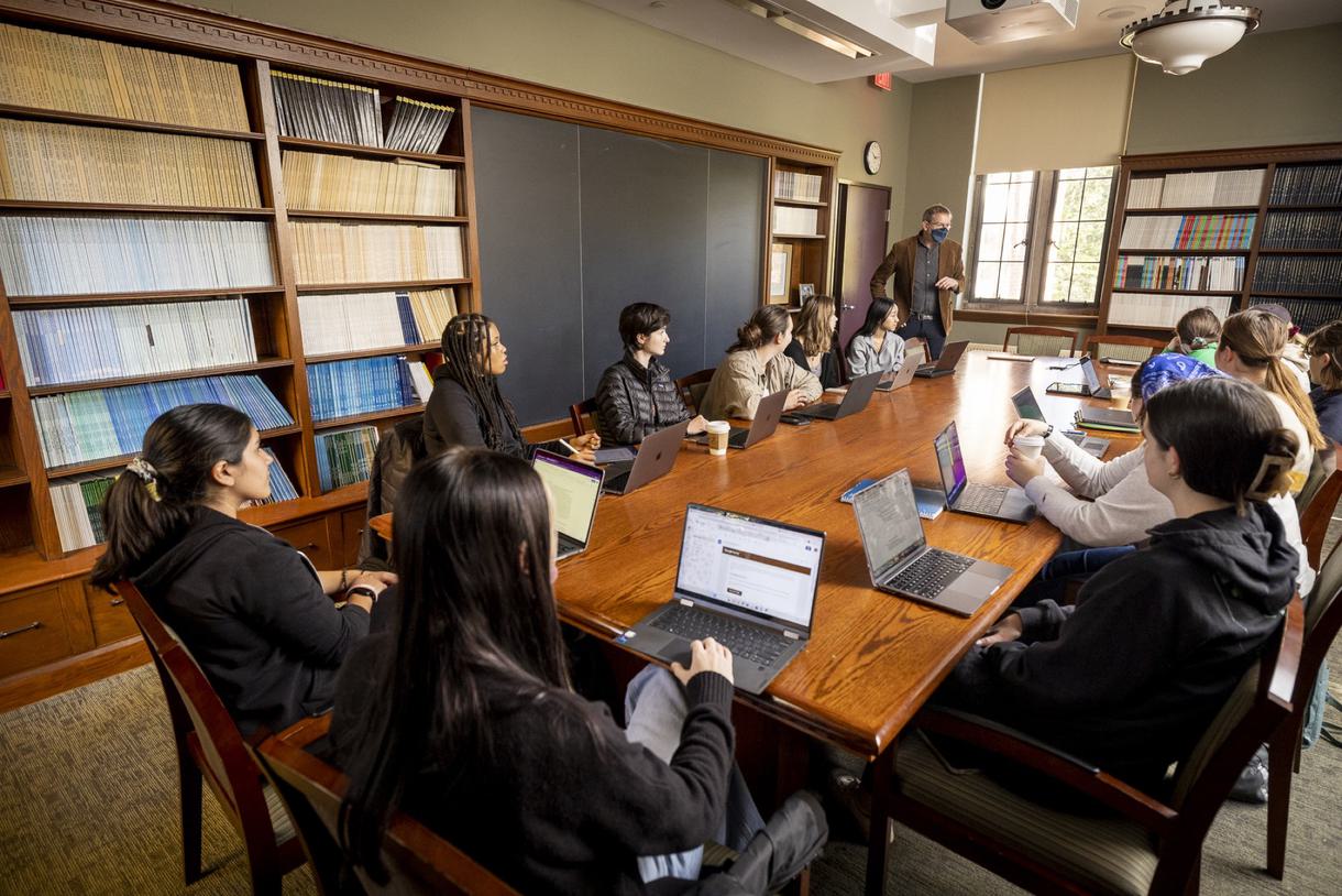 Students sit at a long wooden table in a classroom where books line the shelves.