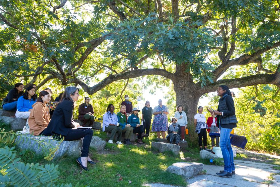 Students, staff and faculty listening to a person who is standing and talking outside. Some are sitting on stone benches and some are standing.