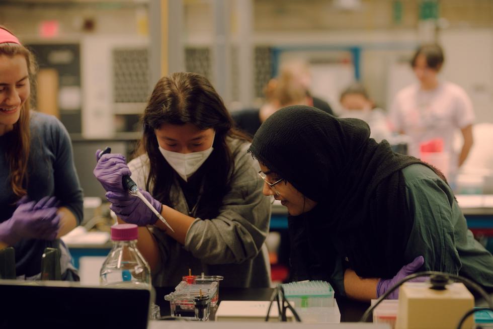Two students observe a third pipetting into a gel in a science lab.