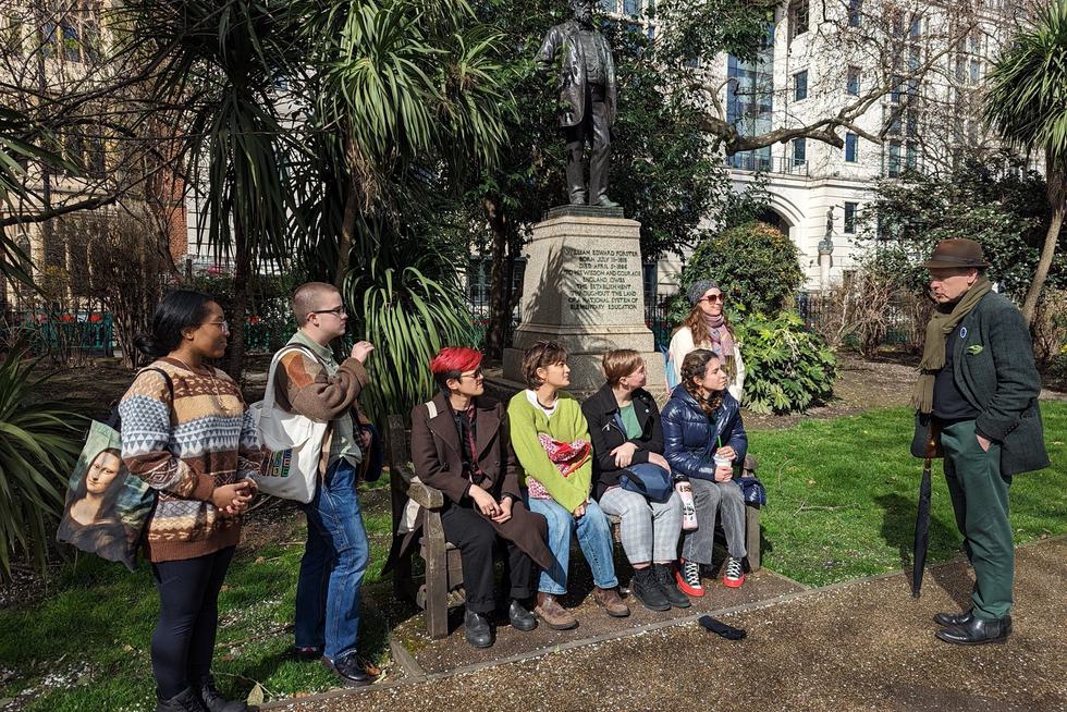 Group of student sit on a bench under a statue of William Edward Forster while listening to a tour guide.