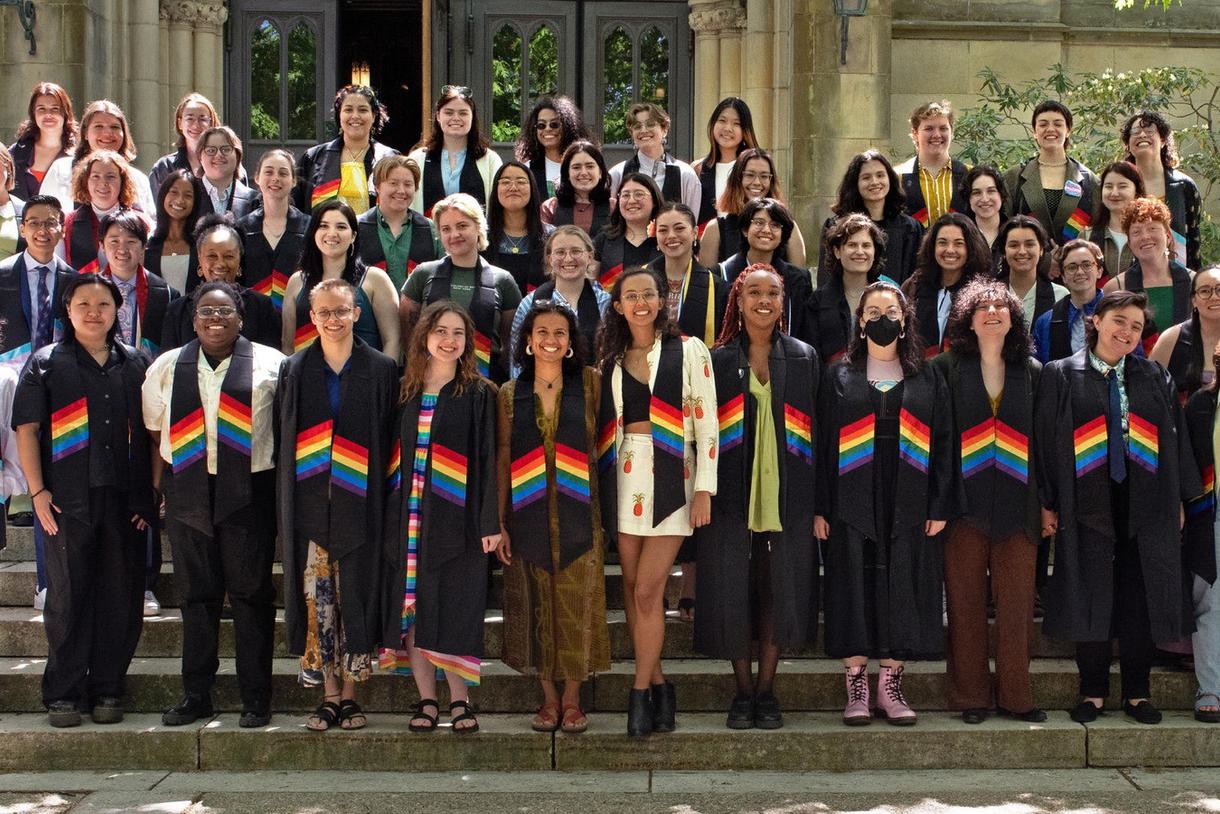 Graduating students pose for a group picture while wearing rainbow stoles. Some are dressed in graduation regalia while others are dressed professionally.