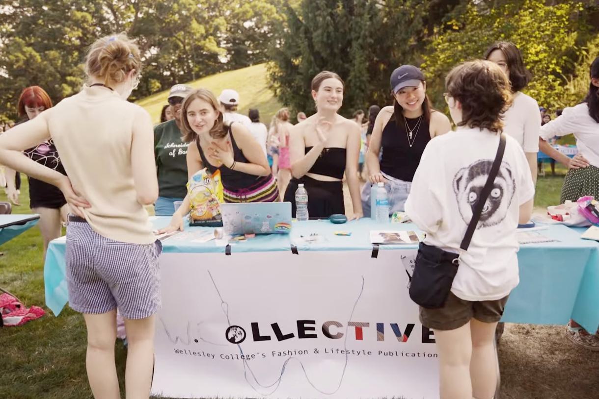 Students representing W. Collective, Wellesley College's Fashion and Lifestyle Publication, stand in front of a table at the annual Student Organization Fair. Two students are facing the table talking with the representatives.
