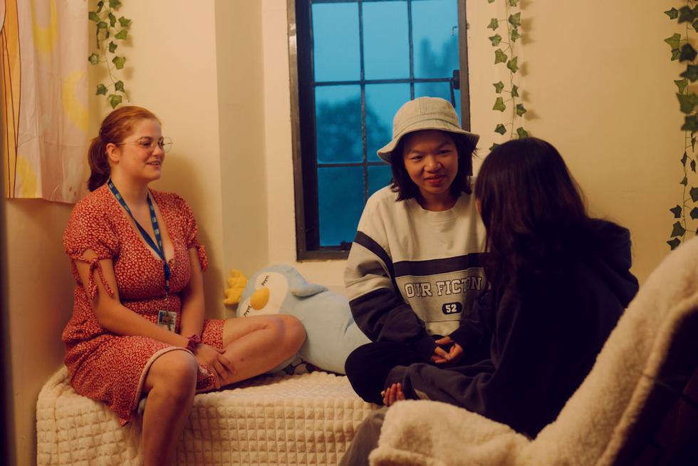 Three students sit on a bed in a dorm room.