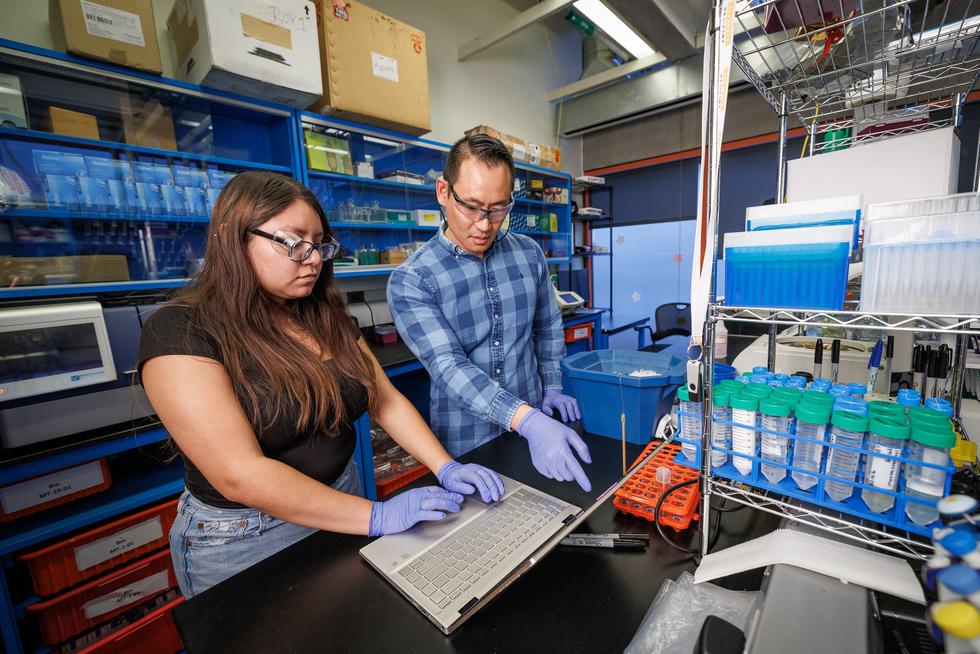 A student wearing goggles and purple gloves holds a pipette and a professor also wearing goggles observes her.
