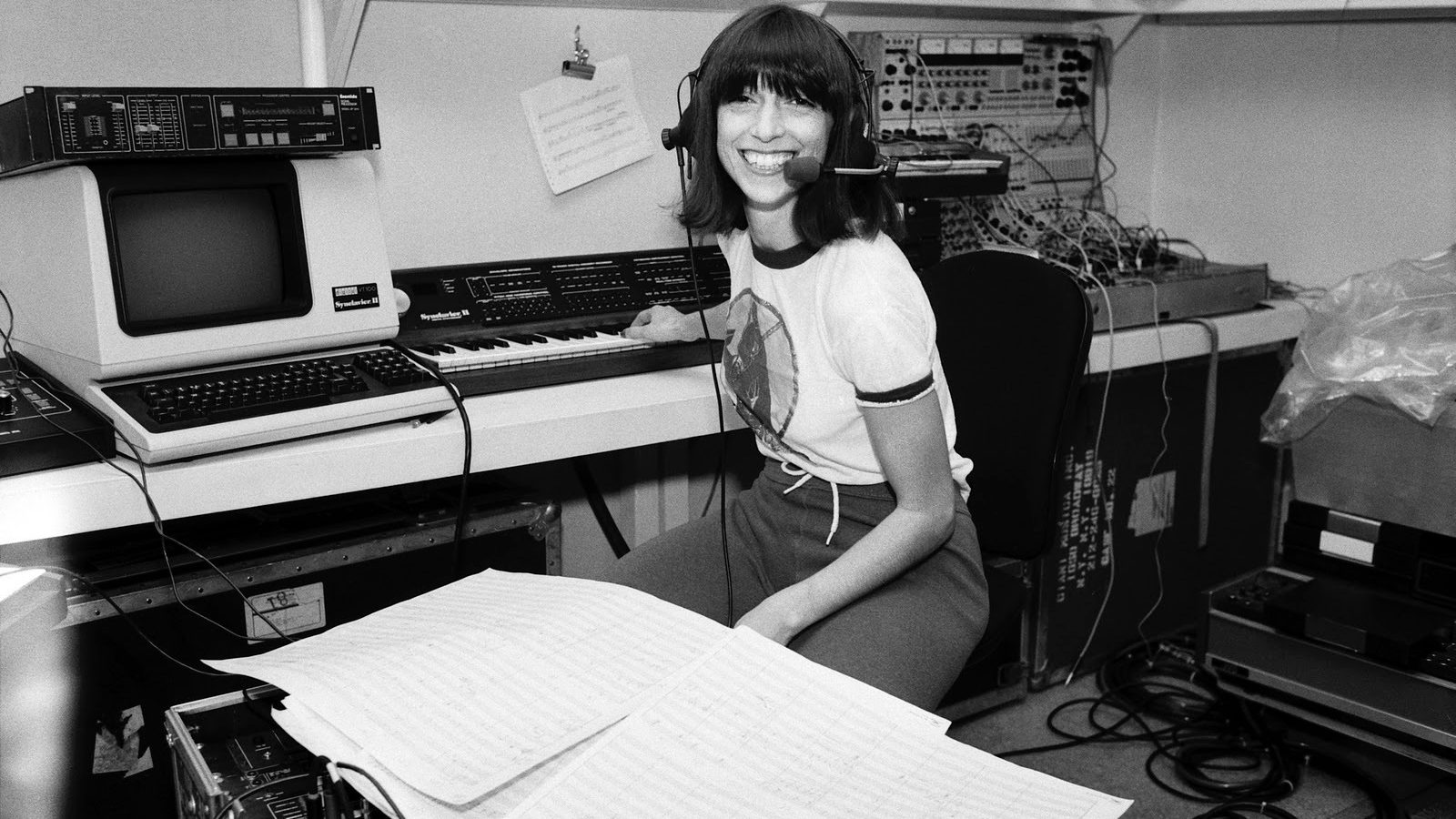 Black and white photo of a young Suzanne Ciani smiling, seated in front of synthesizer