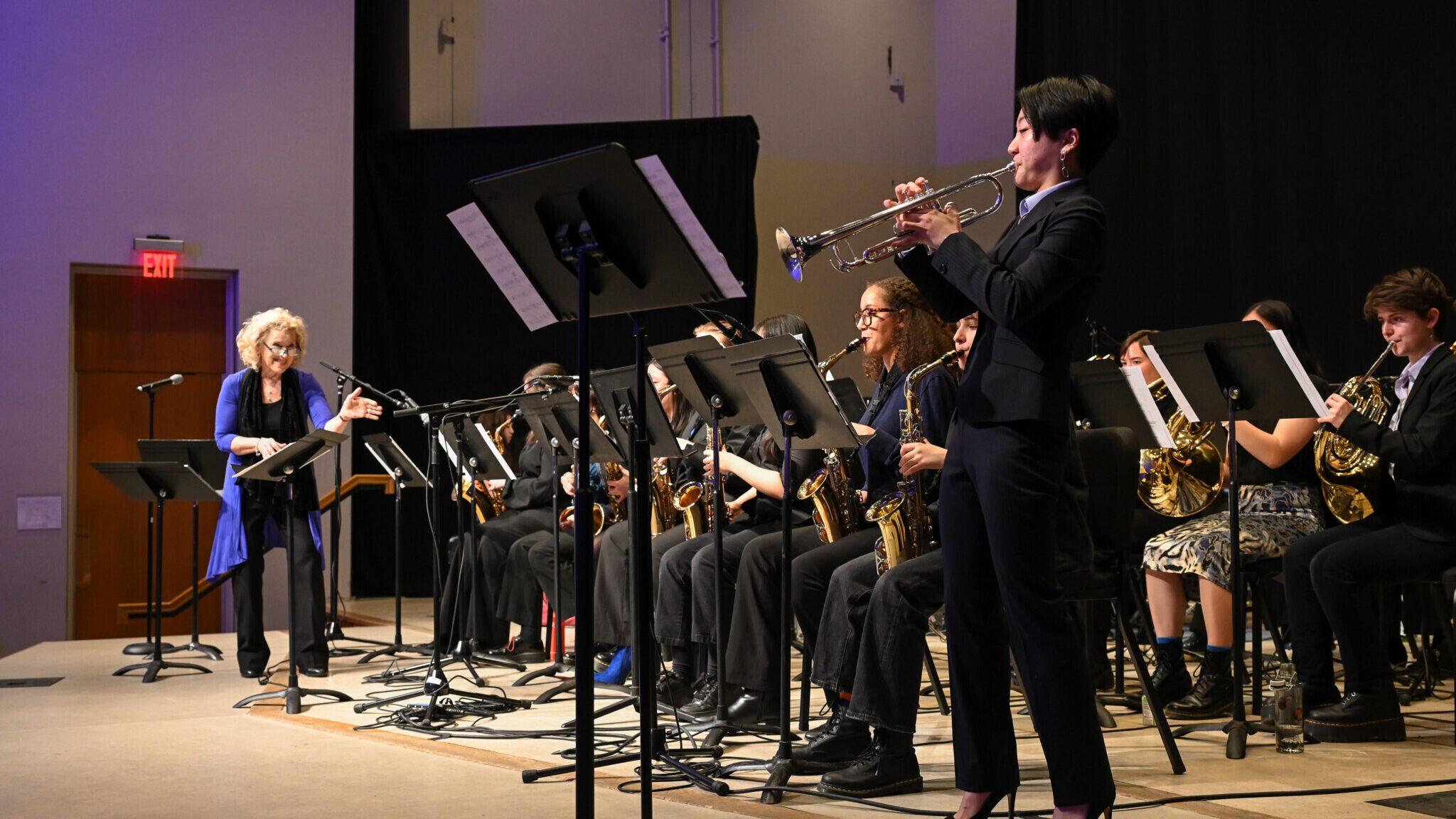 Performers seated and playing on stage, with trumpet player standing and director looking on