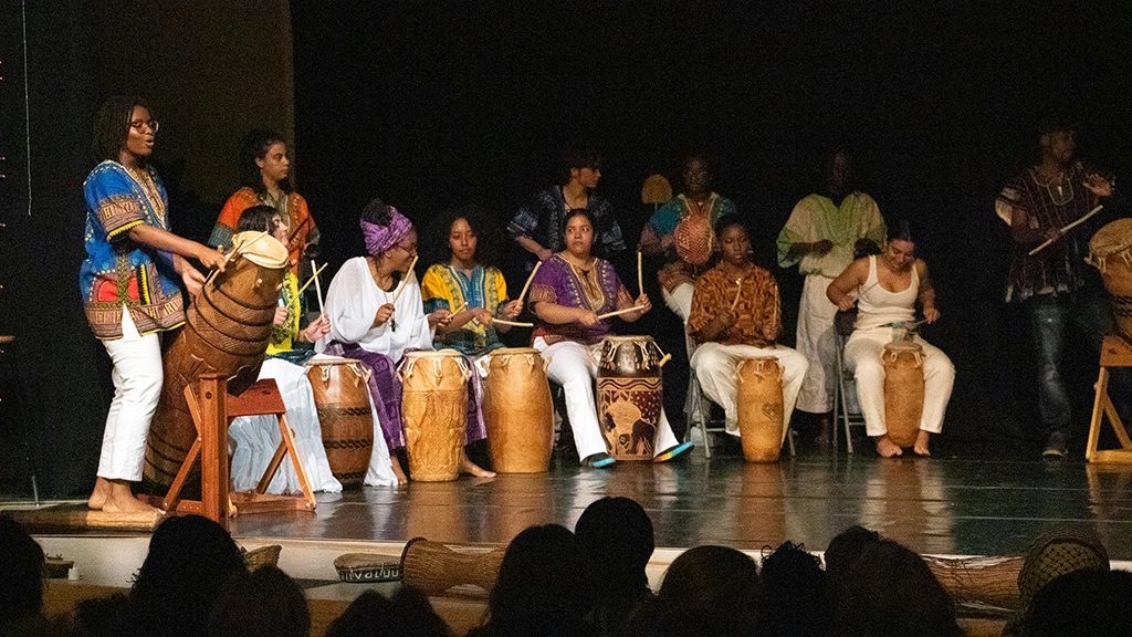 Drummers some standing, some seated, wearing colorful garb, performing onstage.