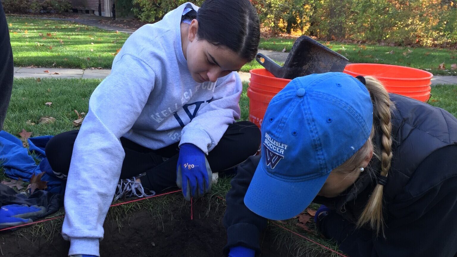 Two students crouched over an excavation site digging with trowels.