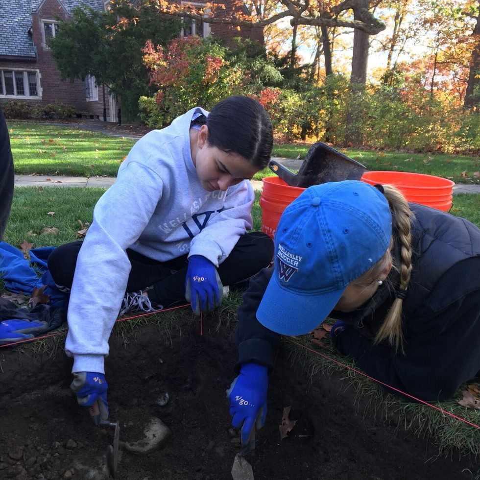 Two students crouched over an excavation site digging with trowels.