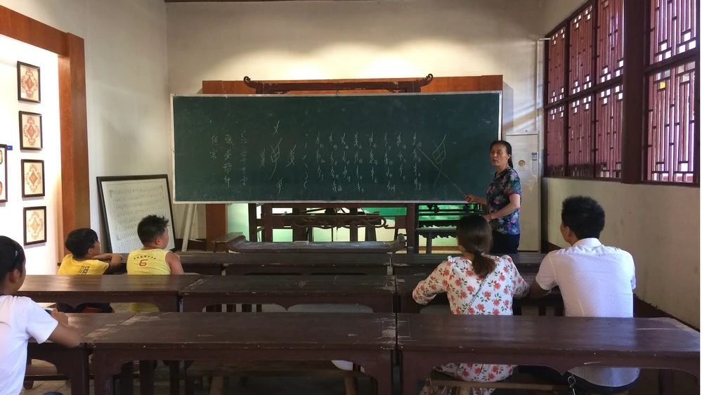 Women at chalkboard with pointer teaching students seated on benches