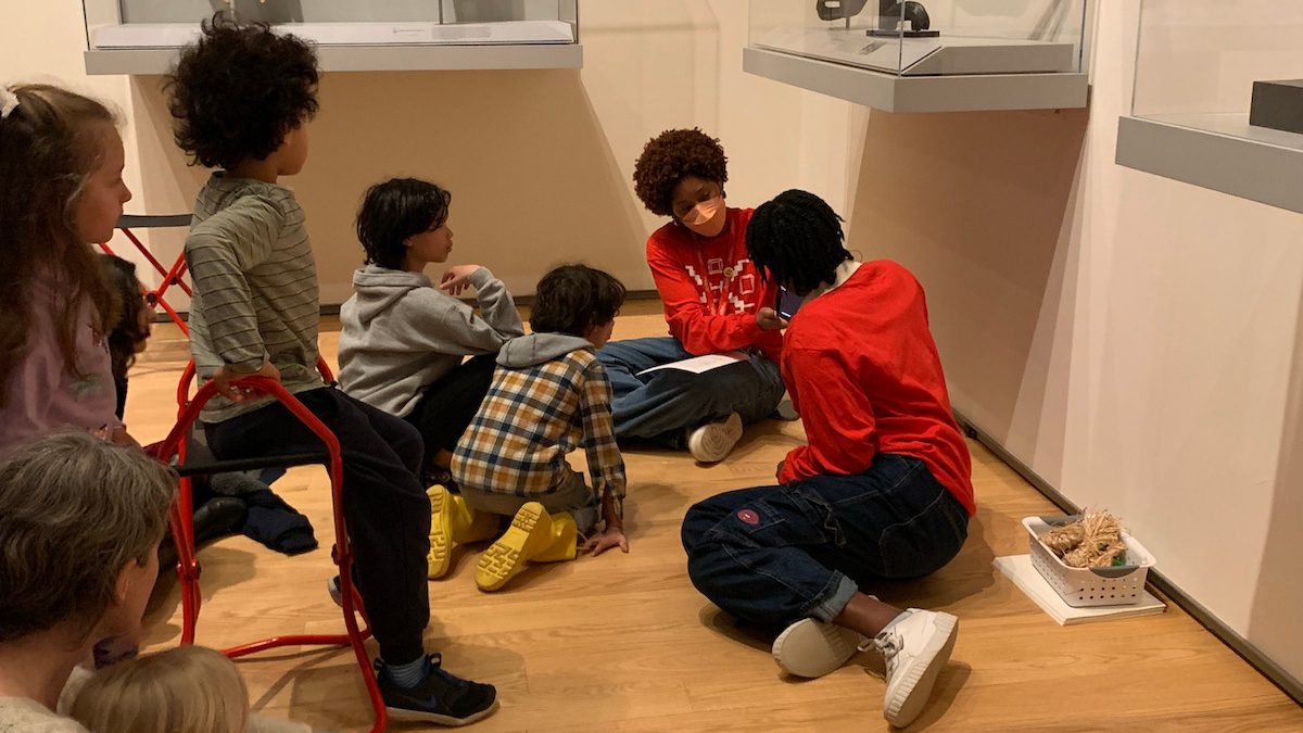 art gallery surrounded by sculptures, student guide engages in teaching art to small children siting on the floor