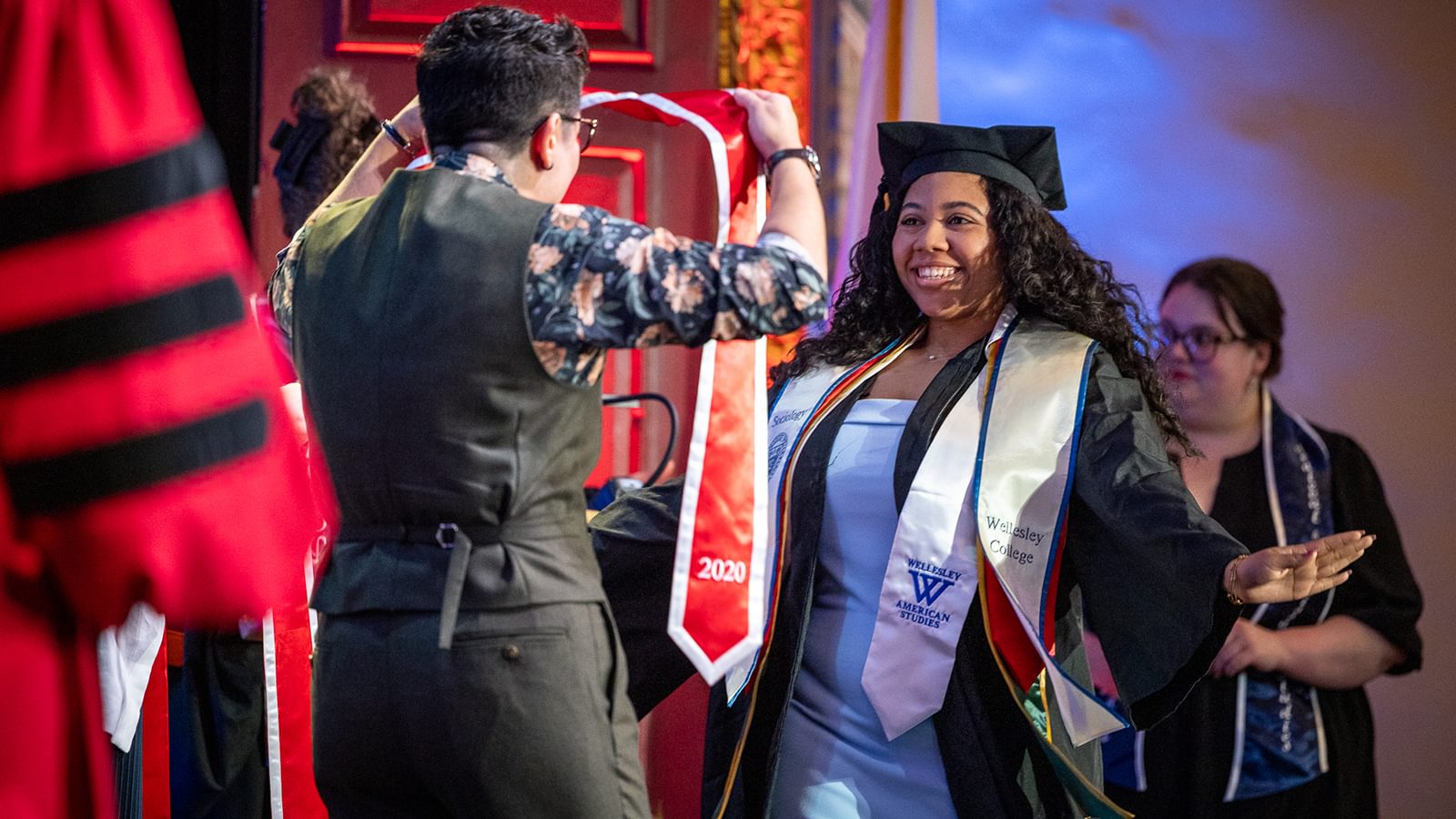 A 2020 lifts her hands and smiles as a Alumnae Board Member prepares to put a red stole around her shoulders.