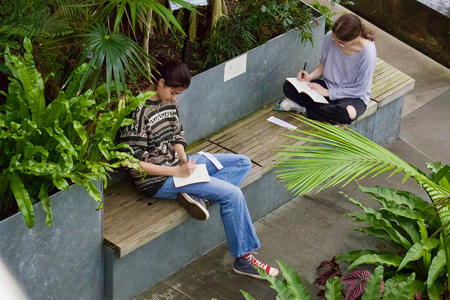 Two student sit inside Global Flora, surrounded by plants and writing in their notebooks.