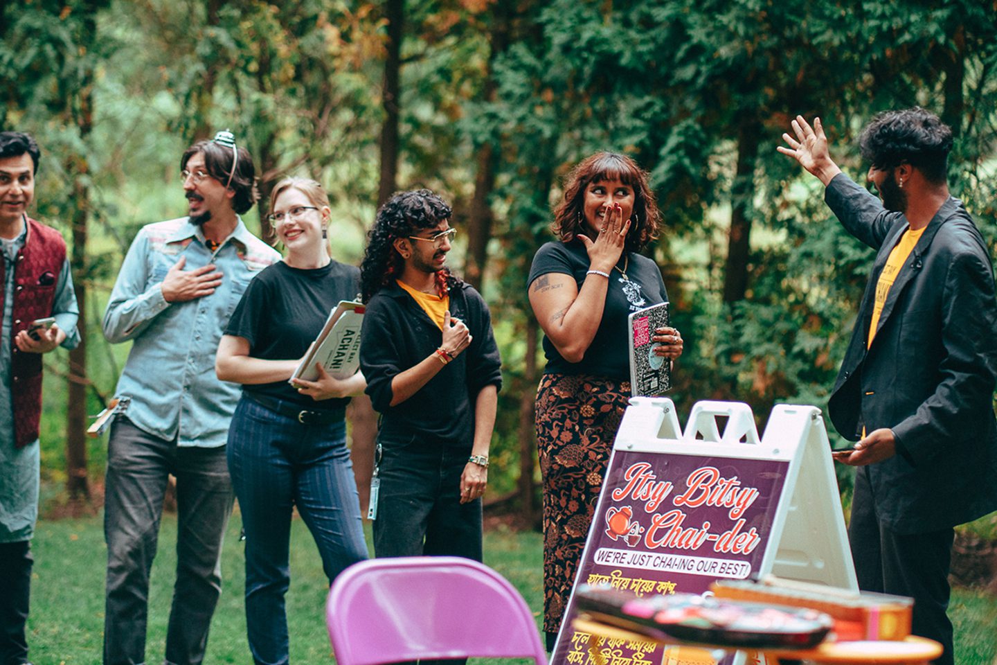 A scene from an outdoor play. There is a line of people and the first person in line is smiling and holding her hand in front of her mouth. She is interacting with a man gesturing with his hand.