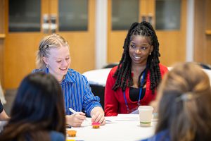 Two students sit at a table. One is smiling while looking down at her hands. The other is smiling while looking at the student.