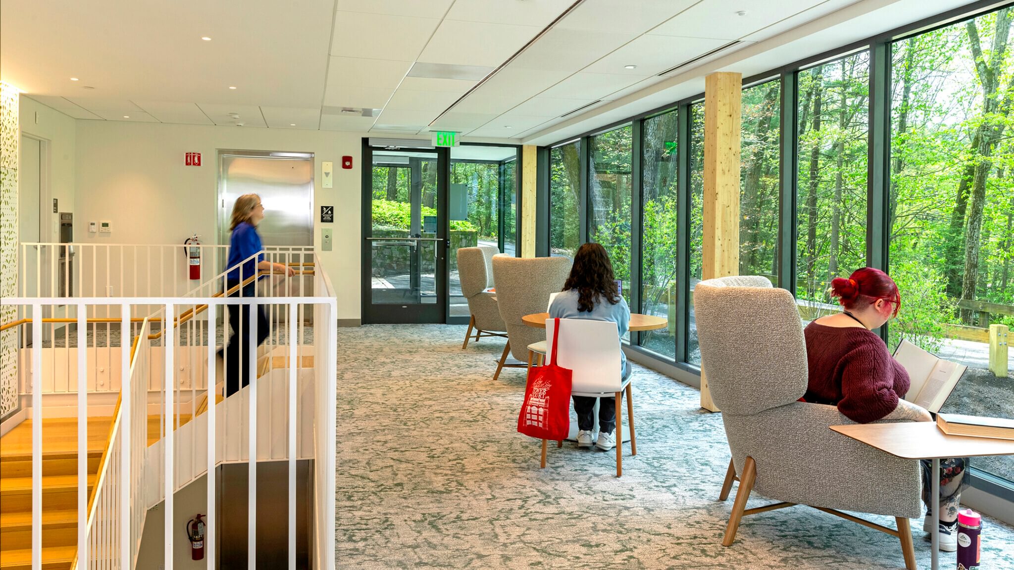 A room with big windows looking out on a lush canopy of trees. One person walks up a staircase while two people sit in chairs looking out the windows.