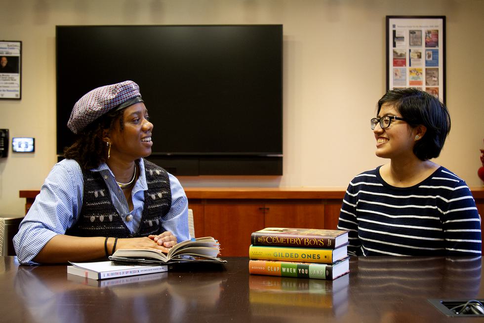 Lillian Goins ’26 and Angelica Delgado ’25 look at each other and chat. They each have books stacked in front of them.