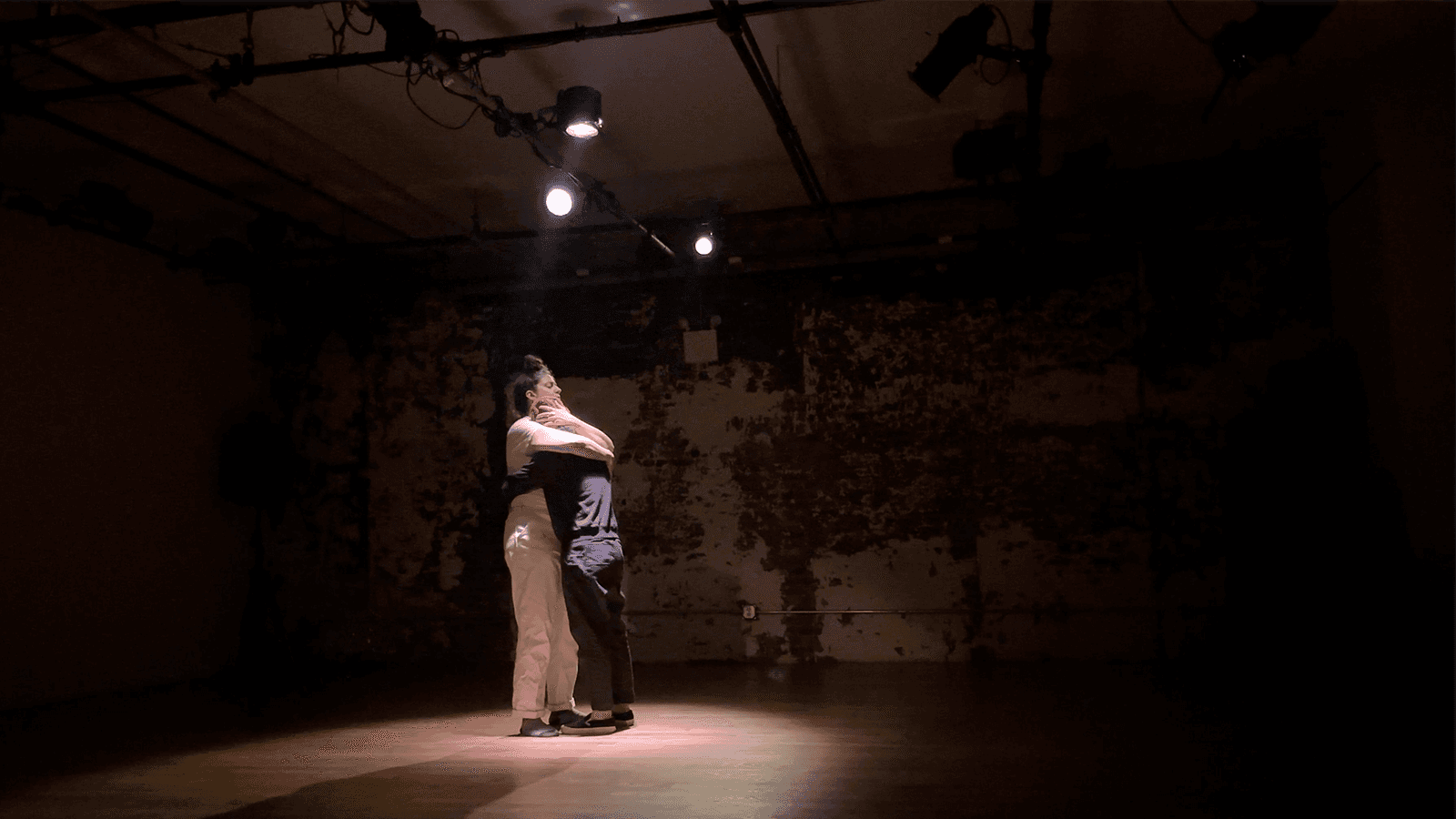 Two people lit by a couple of lights embrace in the middle of a stage that is mostly dark.