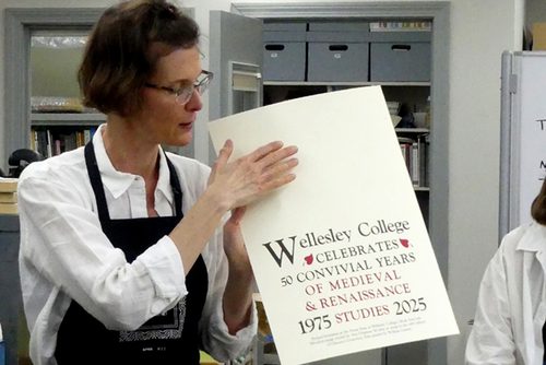 A woman holds a poster that has text on the bottom of the page and is blank at the top. Her hand references the empty space. The poster reads Wellesley College celebrates 50 convivial years of medieval & renaissance studies. 1975, 2025