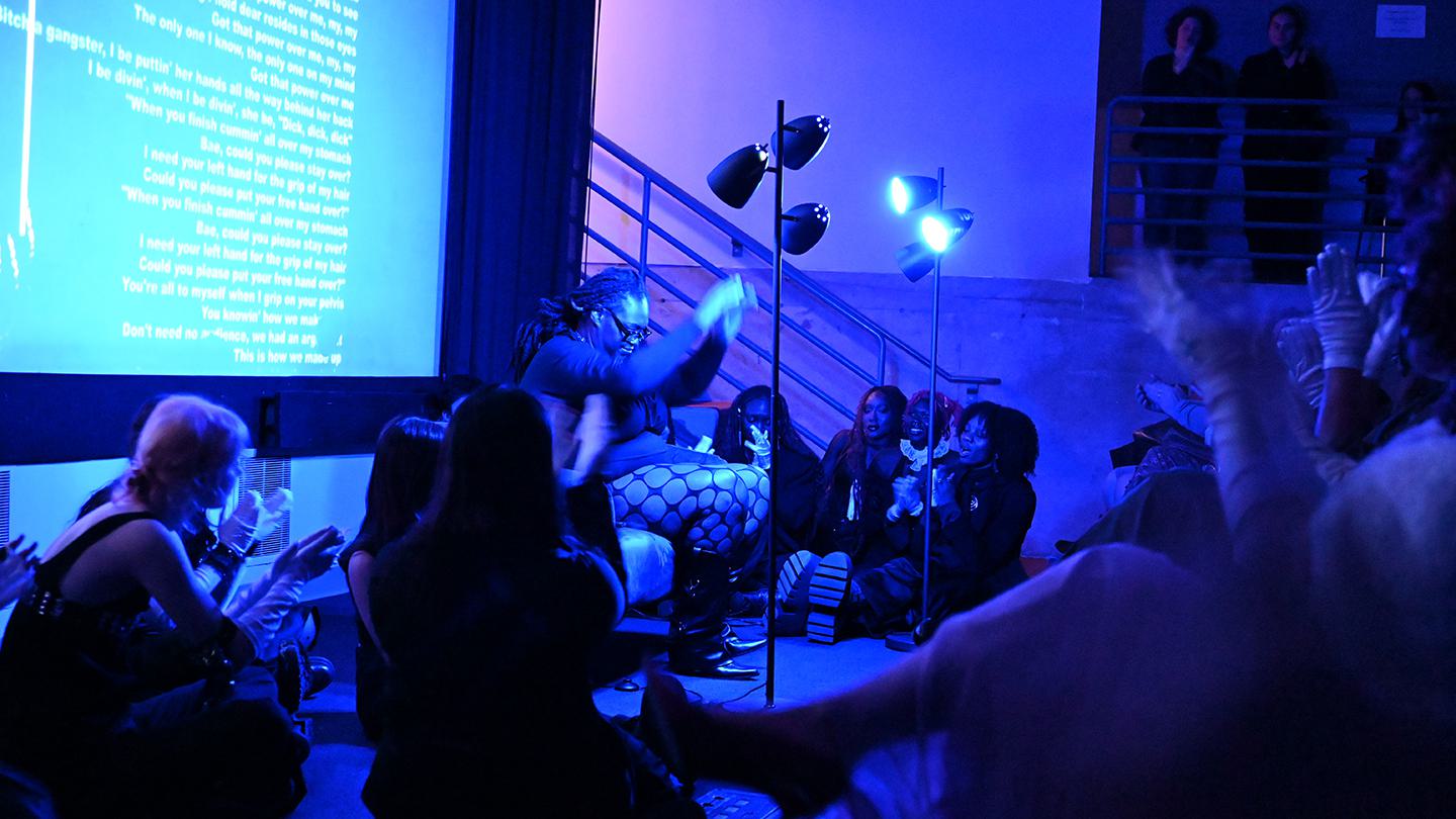 A performance artist is on stage, saturated in blue lighting as students sit on the ground looking up at her.