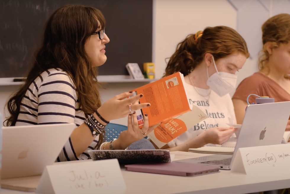 Three students sit at a table looking at a book.