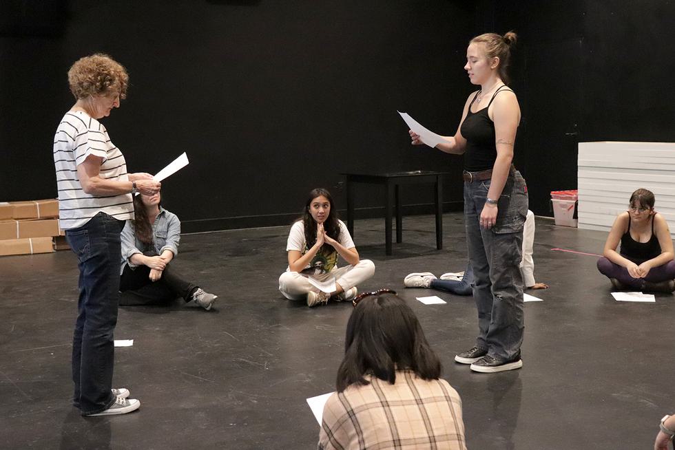 A student and a performer with Actors From The London Stage stand and read lines. Students sit on the ground, watching.