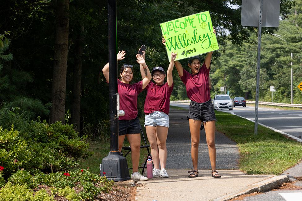 Three students raise their hands in cheer near the entrance of Wellesley. One holds a Welcome to Wellesley sign.