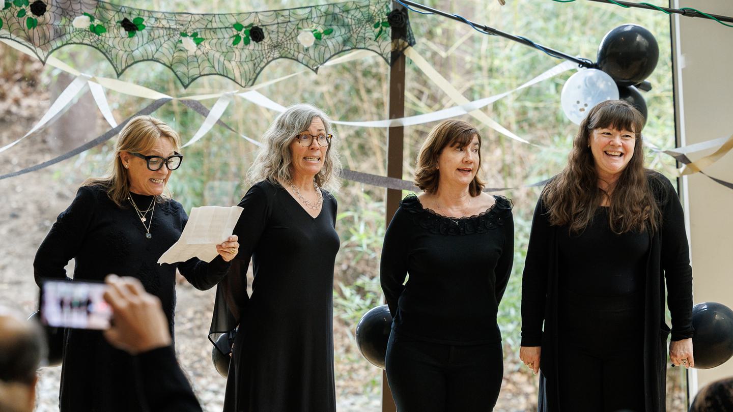 Four women dressed in black stand and sing for an audience.