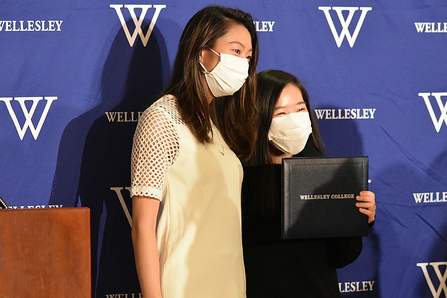 Two students pose for a photo with one student holding up her diploma case that read Wellesley College.